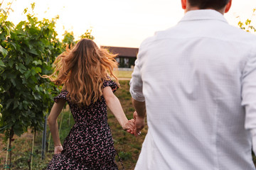 Beautiful young couple man and woman holding hands while walking outdoor together, through vineyard...