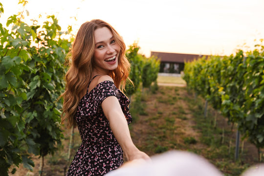 Pretty young woman holding hand of her boyfriend while walking outdoor together, through vineyard on summer day. Follow me concept