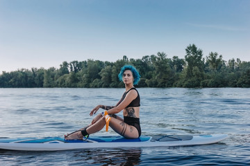 attractive tattooed sportswoman with blue hair relaxing on sup board on river