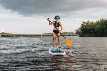 attractive tattooed girl with blue hair paddle boarding on river