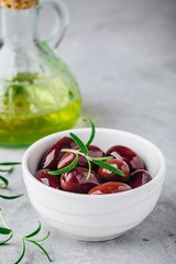Fresh black olives in olive oil in white bowl with rosemary on gray stone background.