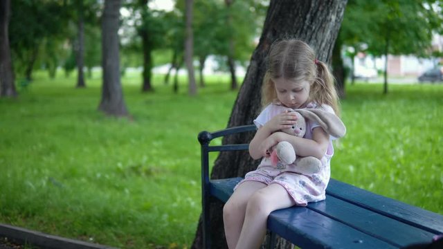A small sad girl in a pink overalls strokes a toy bunny, a child sits on a park bench in a cloudy day.
