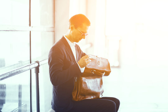 Handsome Businessman Sitting On Bench With Bag On His Lap. Good Looking Man Wearing Elegant Black Suit And White Collar Shirt Taking Look Inside His Leather Bag.