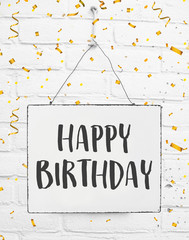 Happy birthday text. Board on white brick background with golden party confetti. Celebrate date of birth.