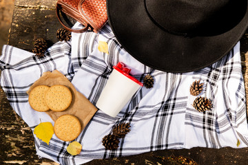 Obraz na płótnie Canvas View of tasty lunch on nature in autumn forest. Flat lay of black hat, scarf, cup with coffee and sweet cookies. Autumn season and picnic concept. Corns on background.