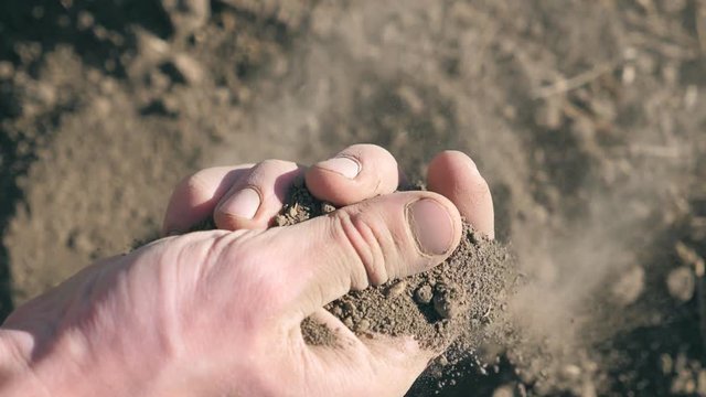 Close up of male farmers hand holding a handful of dry ground and examining it during drought on the field. Young man checks soil fertility on the meadow. Agriculture concept. POV Slow motion