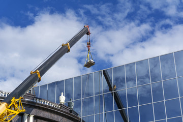 telescopic boom of a construction crane lifts the load against the mirror wall of the building reflecting the sky - Powered by Adobe