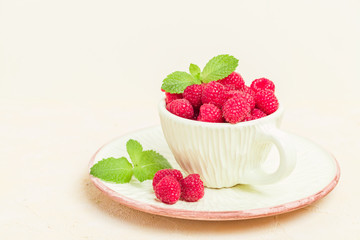 Ripe raspberries with green mint leaves in cup and saucer on pastel yellow background.