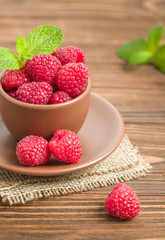 Ripe raspberries with green mint leaves in brown cup and saucer on sackcloth and wooden background.