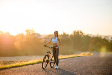 Beautiful happy fit and sporty woman in sportswear standing with bicycle on the countryside road near the field on sunset.