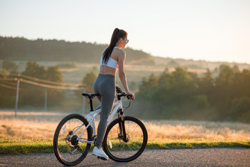 Back view shot of young sporty brunette woman in sportswear riding bicycle outdoors on asphalt countryside road on sunny summer day.