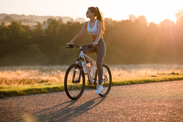 Fototapeta premium Beautiful young sporty brunette woman in stylish sportswear riding on bike on asphalt countryside road on sunset. Summer day
