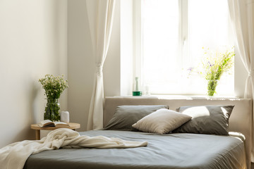 Blanket and pillows on grey bed in bright bedroom interior with flowers and window. Real photo
