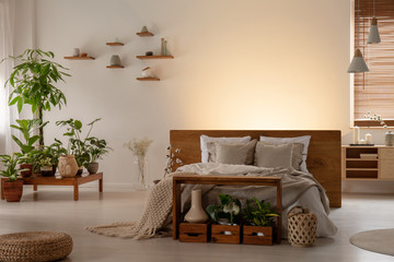 Plants and pouf in dark bedroom interior with light behind wooden bed with headboard. Real photo