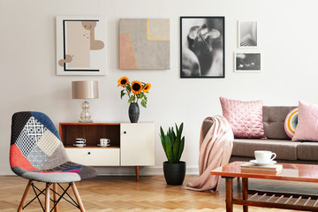 Modern chair and sunflowers on cabinet in flat interior with posters and pink pillows on sofa. Real photo