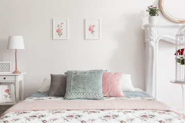 Pastel pillows on bed in feminine white bedroom interior with posters and lamp on cabinet. Real photo - obrazy, fototapety, plakaty