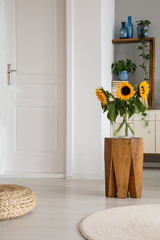 Sunflowers on wooden stool next to pouf in white living room interior with door and rug. Real photo