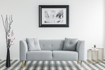 Flowers next to grey sofa with cushions in minimal living room interior with poster. Real photo