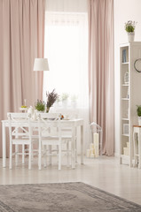 White chairs at table with plants in pastel pink dining room interior with rug and drapes. Real photo