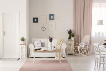 Wooden table on pink rug in white living room interior with rocking chair next to couch. Real photo