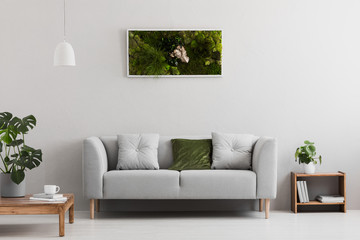 Grey sofa with pillow in real photo of bright sitting room interior with books on wooden shelf, coffee cup on table and garden in frame hanging on the wall