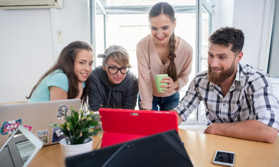 Four co-workers smiling, while watching together a professional business presentation or a funny video on tablet in a modern shared office space for freelancers or young entrepreneurs