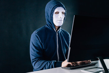 Anonymous and masked hacker under hoodie using computer isolated over dark background - illegal...