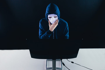 Anonymous and masked hacker under hoodie using computer isolated over dark background - illegal online internet criminal concept.