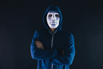 Anonymous masked man under hoodie with arms crossed isolated over dark background - incognito and mysterious criminal on internet activities concept.