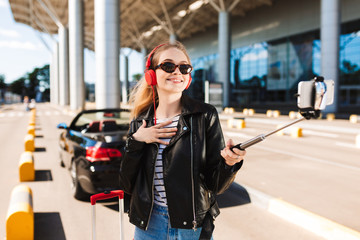 Cheerful girl in sunglasses and headphones taking photo on cellphone with black cabriolet on...