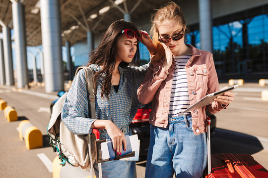 Cool girls in sunglasses holding passport with tickets while thoughtfully using tablet together with airport on background