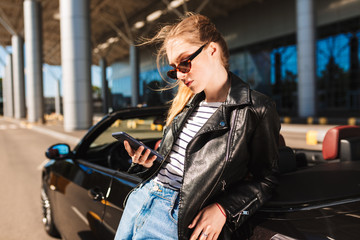 Fototapeta na wymiar Beautiful serious girl in sunglasses and leather jacket leaning on black cabriolet car while thoughtfully using cellphone with airport on background