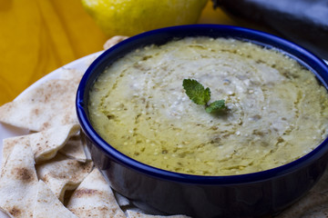 Traditional arabian food, baba ganoush served with syrian bread in a porcelain bowl, garnished with syrian lemon, garlic and eggplant on yellow wooden table
