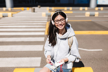 Pretty smiling girl in eyeglasses and headphones joyfully looking in camera with passport and tickets in hands while waiting on road of airport