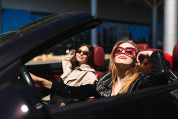 Pretty girl in sunglasses dreamily looking in camera while driving cabriolet car with female friend near on city streets outdoor