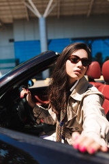 Young beautiful lady in sunglasses and trench coat opening cabriolet car door dreamily looking in...