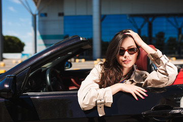 Fototapeta na wymiar Beautiful girl in sunglasses and trench coat leaning on cabriolet car door dreamily looking in camera with airport on background