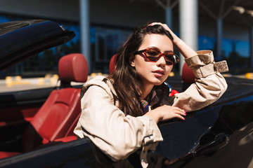 Beautiful girl in sunglasses leaning on cabriolet car door while dreamily looking in camera with...