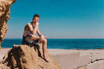 A young guy sits on a rock on the seaside. Near to him the pier, he leaned on his arm and looked at...