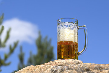 Mug of fresh beer stands on a rock in woods against blue sky