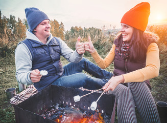 Happy smiling couple roasting marshmallows on campfire and keep their thumbs up - All good. Cheerful man and woman rest at the fire. Date on the nature