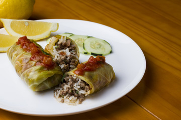 Cigar of cabbage served on white plate accompanied by slices of cucumber, sicilian lemon and tomato on yellow wooden table