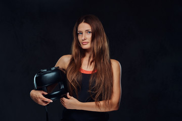 Portrait of a beautiful brunette female boxer in sportswear holds a protective helmet, looking at a camera. Isolated on dark textured background.