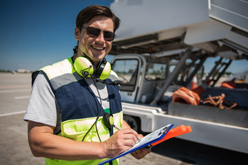 Sunny day. Cheerful man in sunglasses writing information while looking at camera with wide smile. Blue sky, runway and truck on background