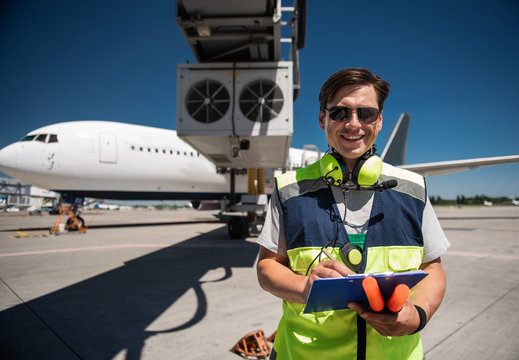 Doing job with joy. Cheerful man in sunglasses noting data and holding signal wands. Passenger plane on background