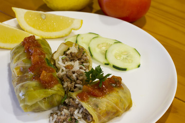 Cigar of cabbage served on white plate accompanied by slices of cucumber, sicilian lemon and tomato on yellow wooden table
