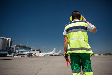 I hear you. Back view of man in signaling vest holding wands and checking the headset. Blue sky,...