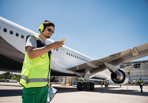 Chatting during work. Smiling man in sunglasses looking at cellphone and holding clipboard. Runway, passenger plane and colleague on background