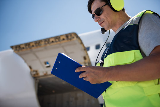 Noting All Details. Low Angle Portrait Of Airport Worker Filling Out Documents. Open Cargo Door On Blurred Background