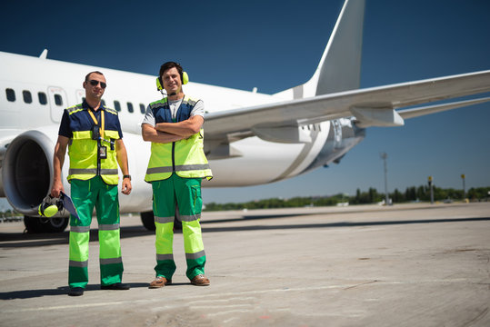 Sunny day at work. Full length portrait of man in sunglasses and smiling colleague with folded arms. Passenger plane, runway and blue sky on background